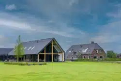 Overzichtsfoto boerenerf transformatie. Schuurwoning met hoge glazen puien en houten constructie, gelegen in een groen open landschap.