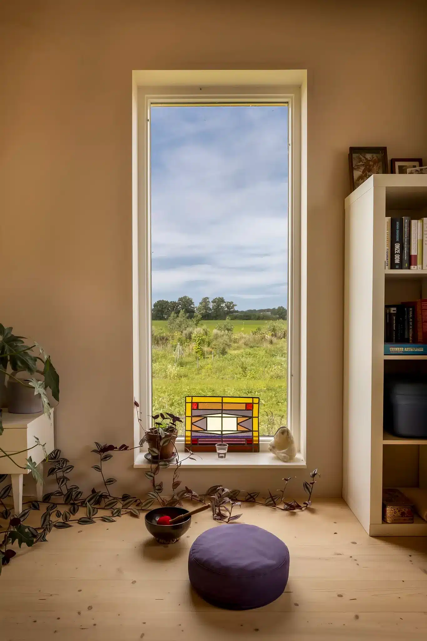 Rustige meditatiehoek met hoog raam, uitzicht op het landschap en klein glas-in-loodpaneel op de vensterbank.