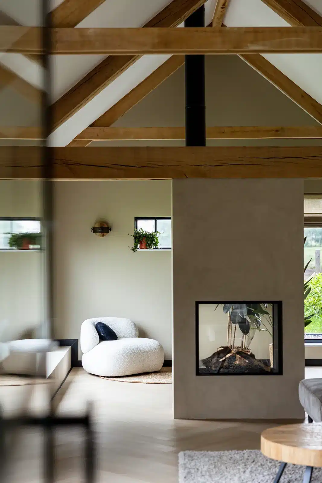 Detailfoto van de zithoek naast de doorkijkhaard, met een witte loungestoel, klein raam met planten en zicht op de houten kapconstructie van de schuurwoning.