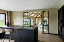 Moderne keuken met donker kookeiland, doorkijk naar de woonruimte met houten spanten en grote harmonicadeuren die uitzicht geven op het omliggende landschap.